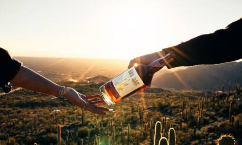 A bottle of whiskey is passed between two people in the desert
